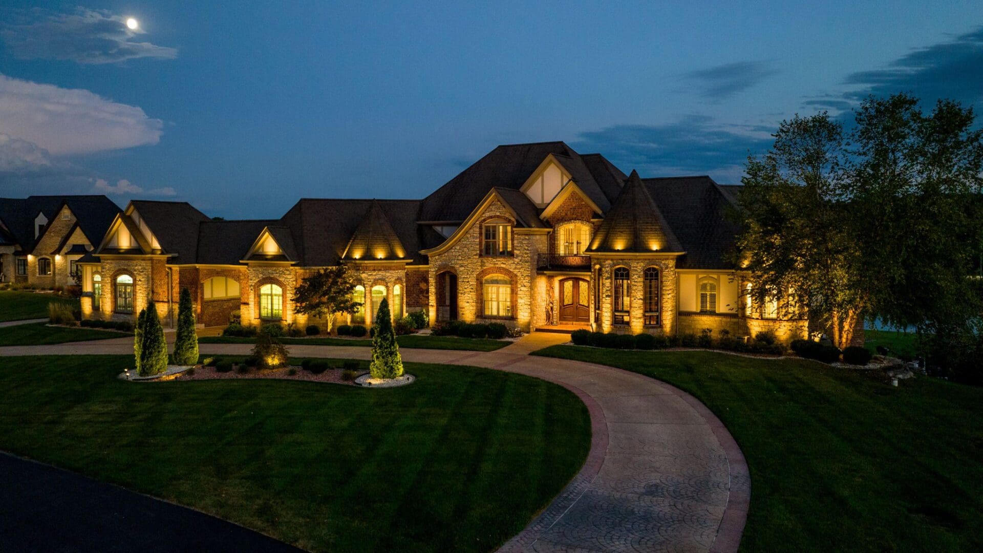 Luxurious mansion illuminated at dusk with a curved driveway and well-manicured lawn.